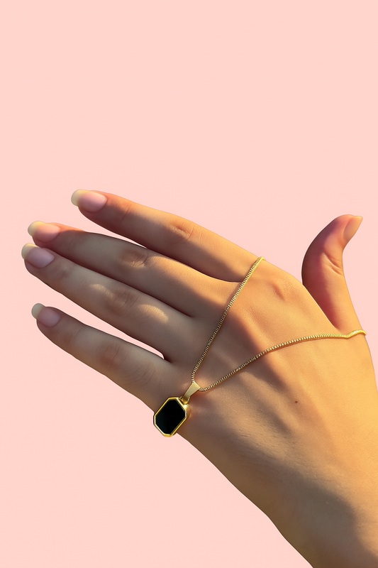 Hand wearing a gold necklace with a black pendant on a pink background