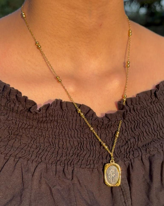 Gold necklace with a pendant on a person wearing a brown top, outdoors.