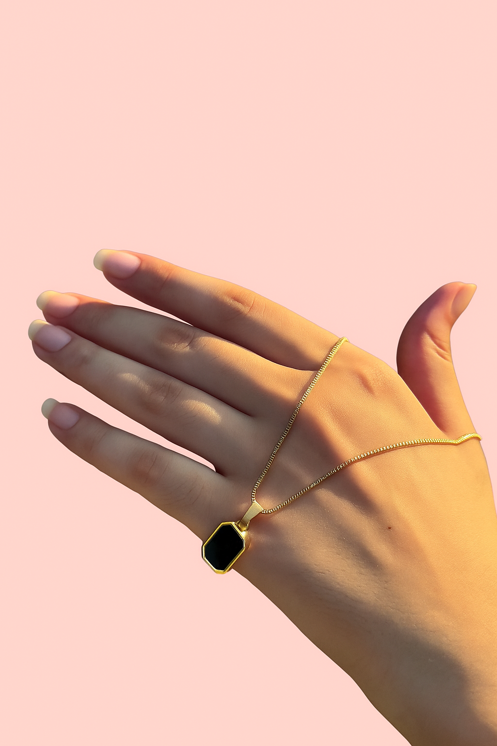 Hand wearing a gold necklace with a black pendant on a pink background